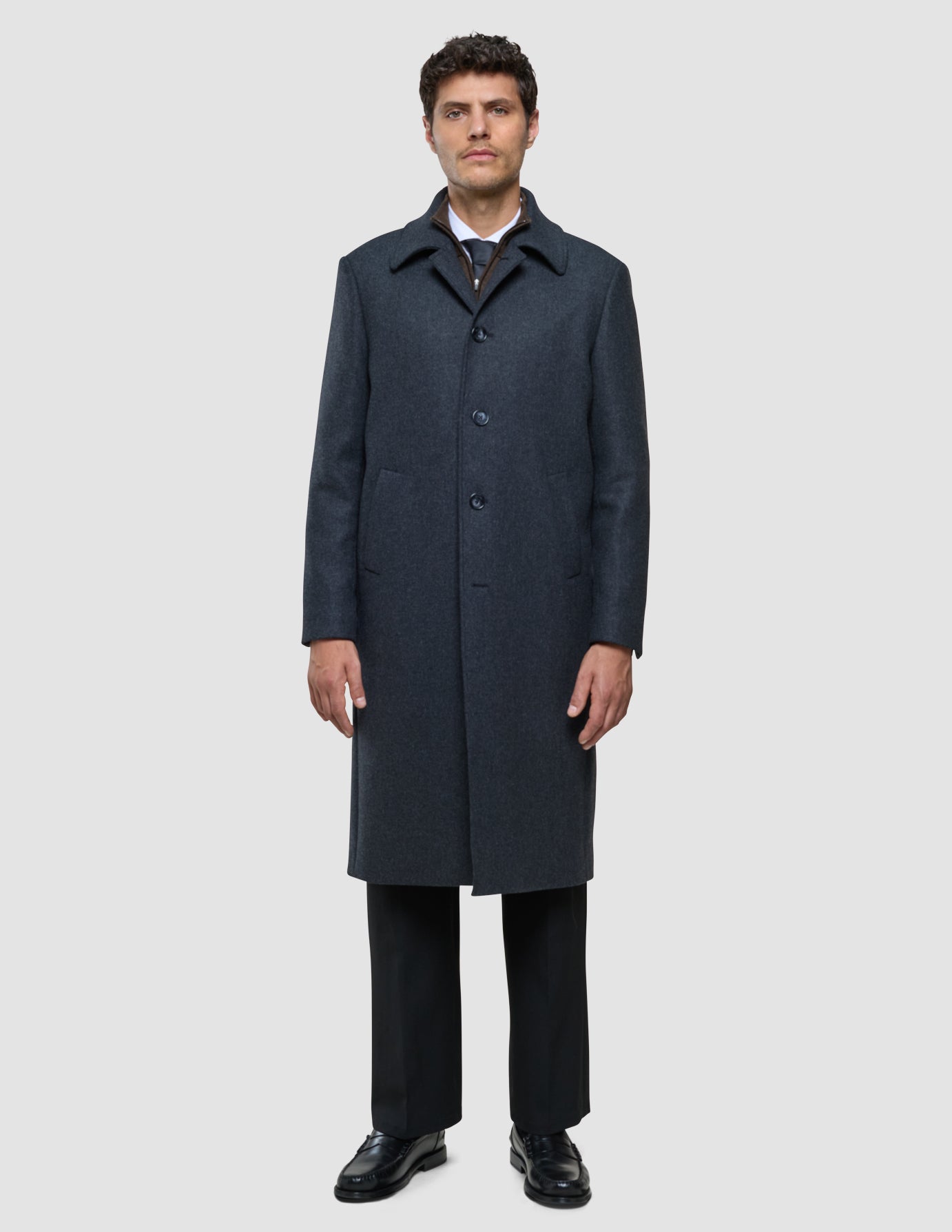 Ein Mann, der einen dunkelgrauen Maxi-Wollmantel über einem Hemd, einer Krawatte und einer schwarzen Hose trägt.